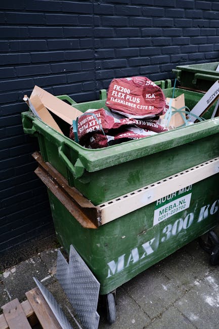 A collection of overflowing rubbish bins and scattered waste bags on a paved area outside a commercial building. The waste includes black, red, and grey bins, with some lids open, revealing mixed pape