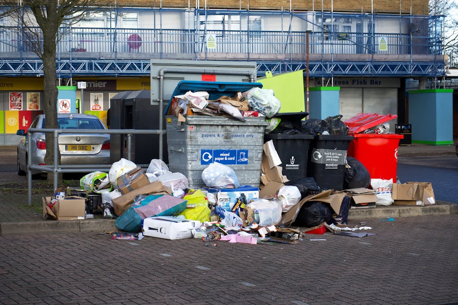 A large pile of mixed waste is overflowing from several black, gray, and red rubbish bins situated on a paved sidewalk area. The waste includes crumpled cardboard boxes, paper bags, plastic packaging,