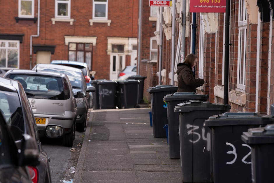 An outdoor scene featuring a worker in a high-visibility yellow and orange vest operating a large red rubbish collection vehicle on a paved street. The vehicle's rear hopper is open, revealing interna